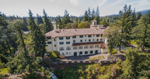 Aerial view of a large, historic building surrounded by trees and a small stream, with a clear blue sky above.