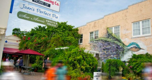 A vibrant mural of a bird adorns a building surrounded by greenery, with people enjoying outdoor dining nearby.