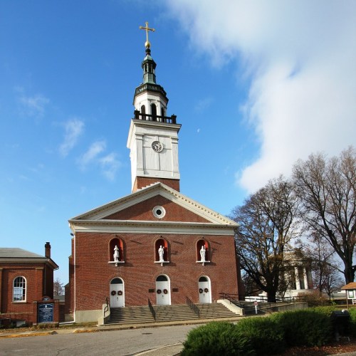 Historic church with a tall clock tower and cross, surrounded by trees under a blue sky with scattered clouds.