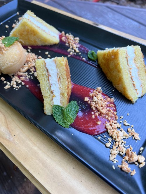 A black plate with layered cake slices, raspberry sauce, granola, and a scoop of ice cream, garnished with mint leaves.