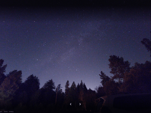 A starry night sky with a faint Milky Way, framed by silhouettes of trees and a vehicle in the foreground.