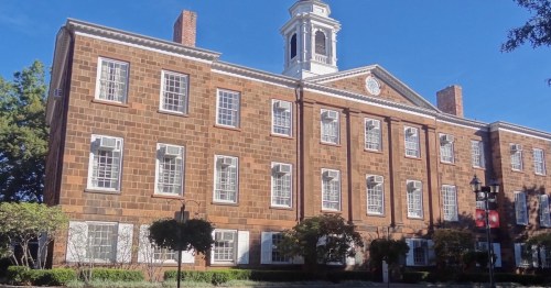 Historic brick building with a clock tower, surrounded by trees and blue sky.