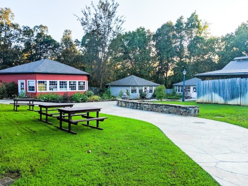 A serene outdoor area with picnic tables, stone pathways, and colorful buildings surrounded by trees.