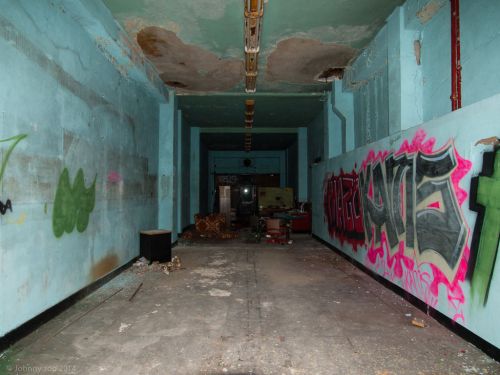 An empty, abandoned room with peeling blue walls and colorful graffiti, featuring debris on the floor and dim lighting.