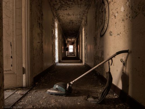 An abandoned hallway with peeling walls and a vacuum cleaner lying on the floor, illuminated by light at the end.