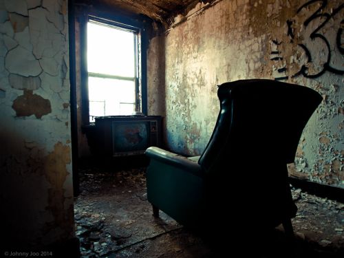 An abandoned room with a cracked wall, a worn-out armchair, and an old television set by a window.