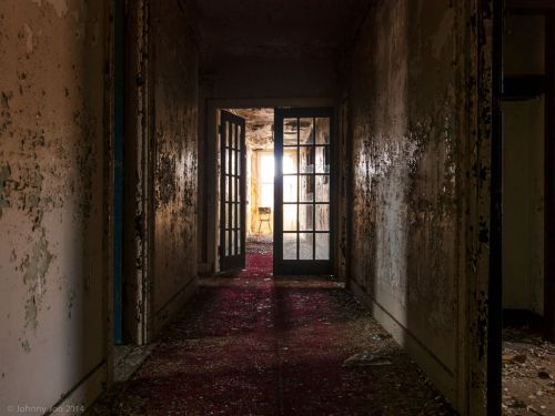 A dimly lit, abandoned hallway with peeling walls and a red carpet leading to a bright doorway at the end.