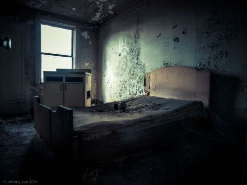 An abandoned, dilapidated bedroom with a worn bed, peeling walls, and dim lighting.