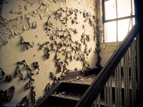 Peeling paint on a wall beside a staircase, with sunlight filtering through a window, creating a moody atmosphere.