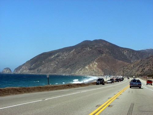 A scenic coastal road with cars, mountains in the background, and waves crashing on the shore under a clear blue sky.