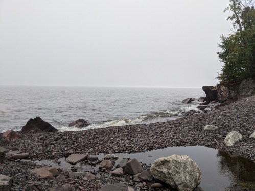 A misty shoreline with smooth stones and gentle waves lapping at the rocky beach under a gray sky.