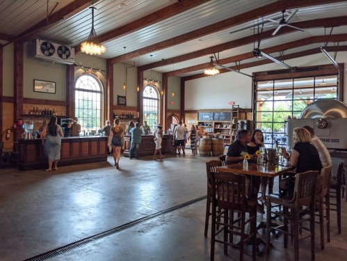 A spacious brewery interior with a bar, patrons enjoying drinks, and large windows letting in natural light.
