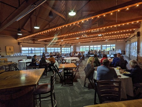 A spacious restaurant interior with string lights, tables, and patrons enjoying their meals. Large windows provide natural light.