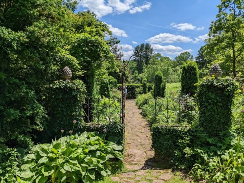 A lush garden path leads through green foliage and ornate gates under a bright blue sky with fluffy clouds.