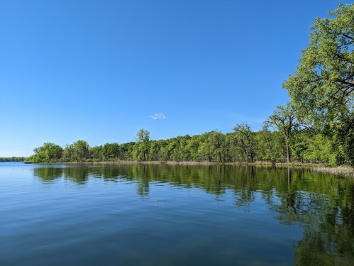 A calm lake reflecting a clear blue sky and lush green trees along the shoreline.