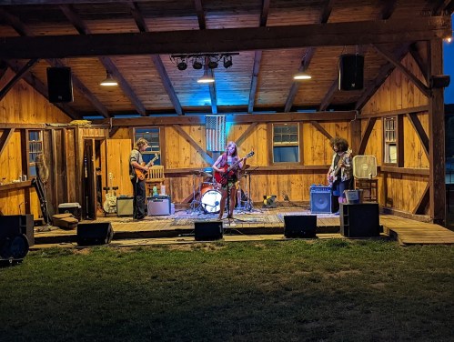 A band performs on stage in a wooden pavilion, with instruments and amplifiers set up under warm lighting.