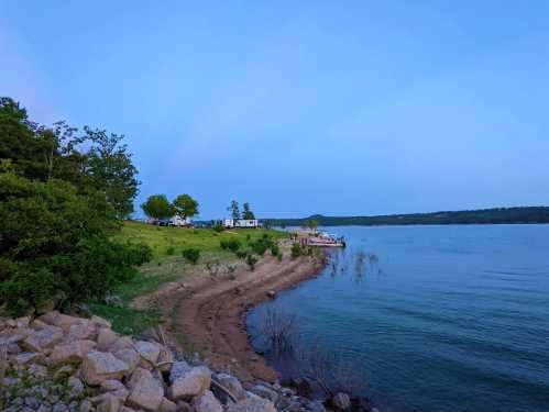 A serene lakeside view at dusk, featuring a winding shore, green trees, and distant campers.