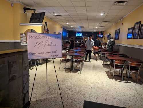 A restaurant interior with tables and chairs, featuring a welcome sign and a few patrons in the background.