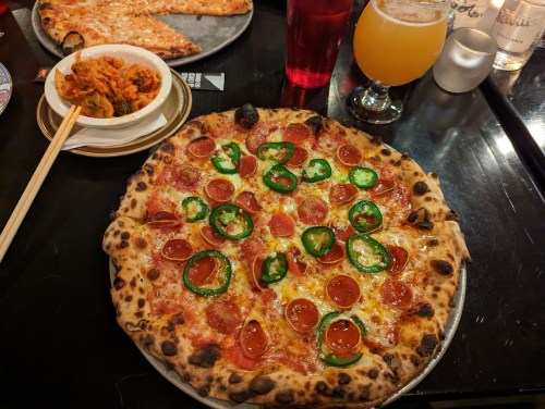 A delicious pizza topped with pepperoni and jalapeños, alongside a plate of spicy chicken and a drink.