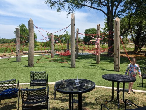 Children play on a colorful climbing structure in a park, surrounded by green grass and trees under a clear blue sky.