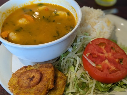 A bowl of soup with shrimp and greens, served with rice, lettuce, tomato, and fried plantains on the side.