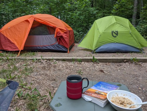 Two tents in a forested campsite, with a coffee mug and a book on a table in the foreground.