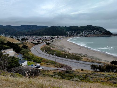 A coastal view featuring a winding road, beach, and hills under a cloudy sky.