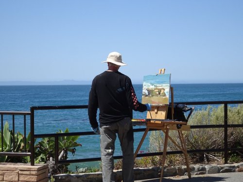 An artist paints a seascape on an easel by the ocean, with a clear blue sky and greenery nearby.