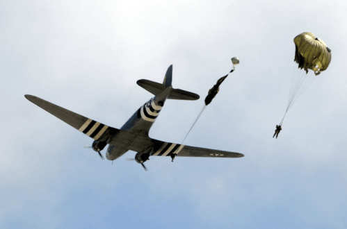A vintage aircraft with striped wings flies overhead as two parachutists descend from it against a cloudy sky.