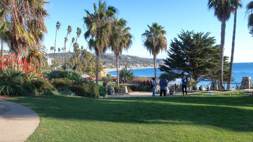 A scenic coastal view with palm trees, lush greenery, and people walking along a path by the ocean.
