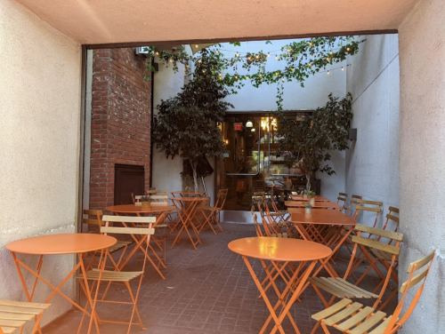 A cozy outdoor seating area with orange tables and wooden chairs, surrounded by greenery and brick walls.