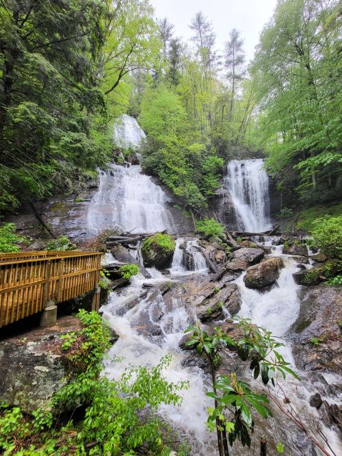A scenic view of cascading waterfalls surrounded by lush green trees and rocky terrain, with a wooden walkway in the foreground.