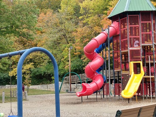 A playground with a red slide, yellow slide, swings, and colorful trees in the background during autumn.