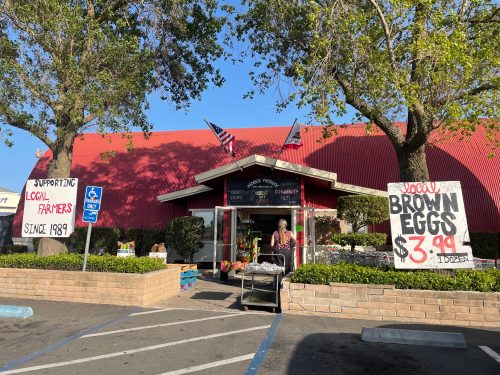 A farm stand with a red roof, signs for local brown eggs, and trees in front, promoting local farmers since 1989.