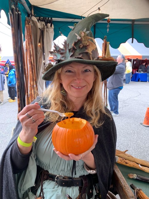 A woman in a witch hat holds a carved pumpkin, smiling while eating from it at a festive outdoor market.