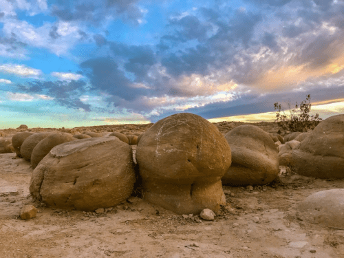 Boulders of varying sizes on a desert landscape under a colorful sky with clouds at sunset.