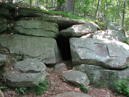 A rocky entrance to a cave surrounded by trees and foliage in a wooded area.
