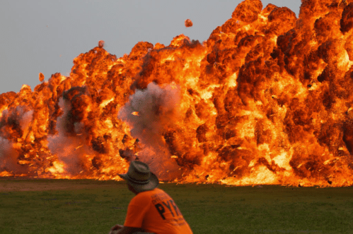 A person in an orange shirt watches a massive explosion, with flames and smoke rising dramatically in the background.