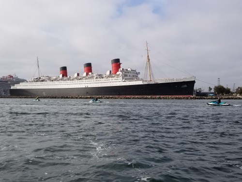 A large historic ship with red and black smokestacks docked near the shore, with jet skis in the foreground.
