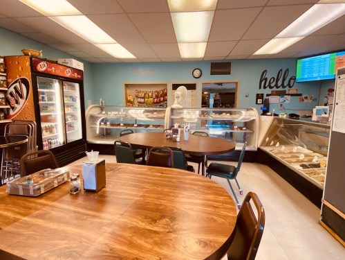 Cozy café interior with a wooden table, display cases of pastries, a soda fridge, and a welcoming "hello" sign.