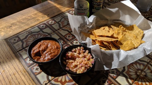 A basket of tortilla chips with two bowls of salsa and pico de gallo on a patterned table.