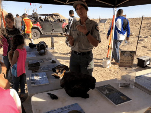 A park ranger explains exhibits at an outdoor event, with visitors and off-road vehicles in the background.