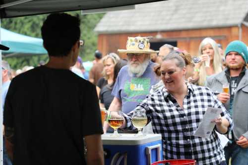 A woman in a checkered shirt serves drinks at an outdoor event, with a crowd and a man in a straw hat in the background.