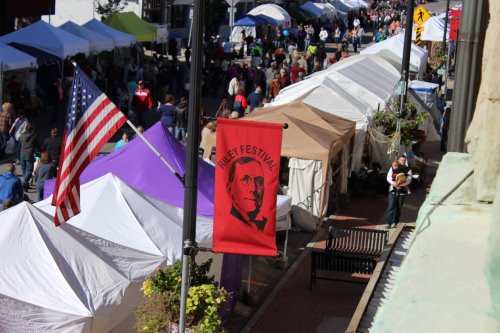 A bustling street market with tents, a festival banner, and an American flag, filled with people enjoying the event.