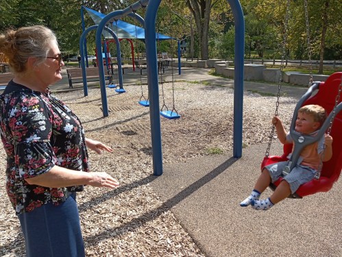 A woman watches a young child happily swinging at a playground surrounded by trees and play equipment.