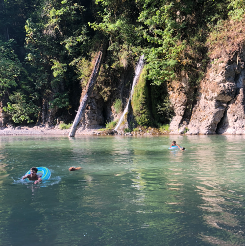 Two people swim in a clear river surrounded by lush greenery and rocky cliffs, with a small waterfall in the background.
