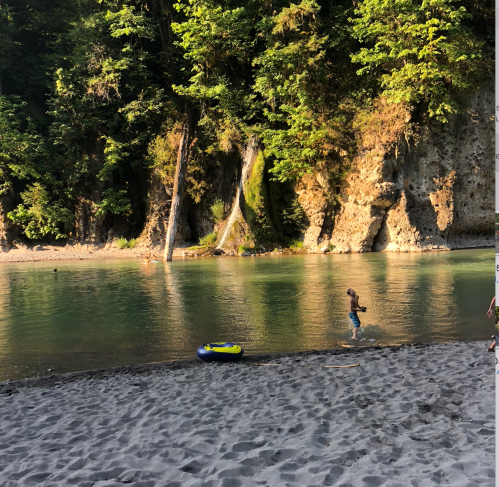 A child runs along a sandy riverbank, surrounded by lush greenery and rocky cliffs. A small inflatable raft is nearby.