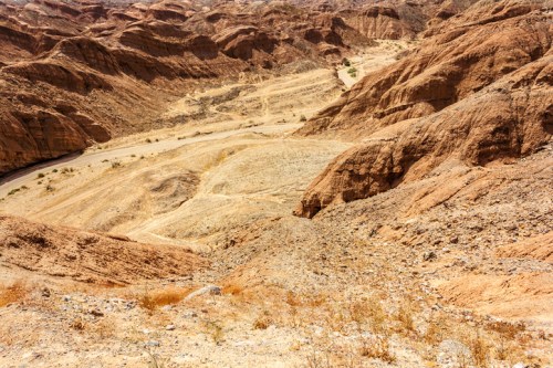A vast, arid landscape of rugged, brown hills and valleys under a clear blue sky.