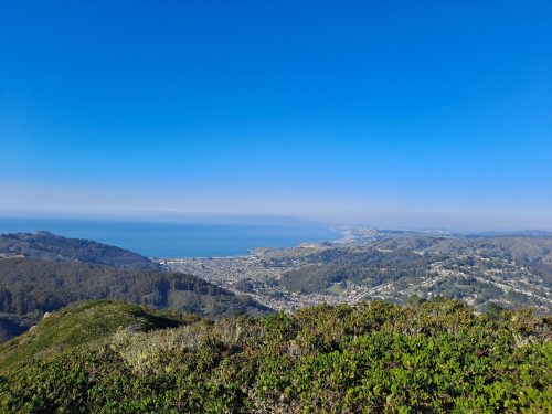 A panoramic view of a coastal town with blue skies and ocean, surrounded by green hills and vegetation.