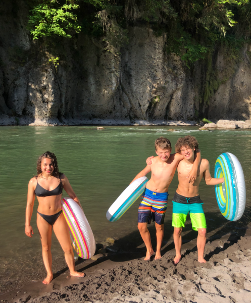 Three young people stand by a river, holding colorful inner tubes, with lush greenery and rocky cliffs in the background.